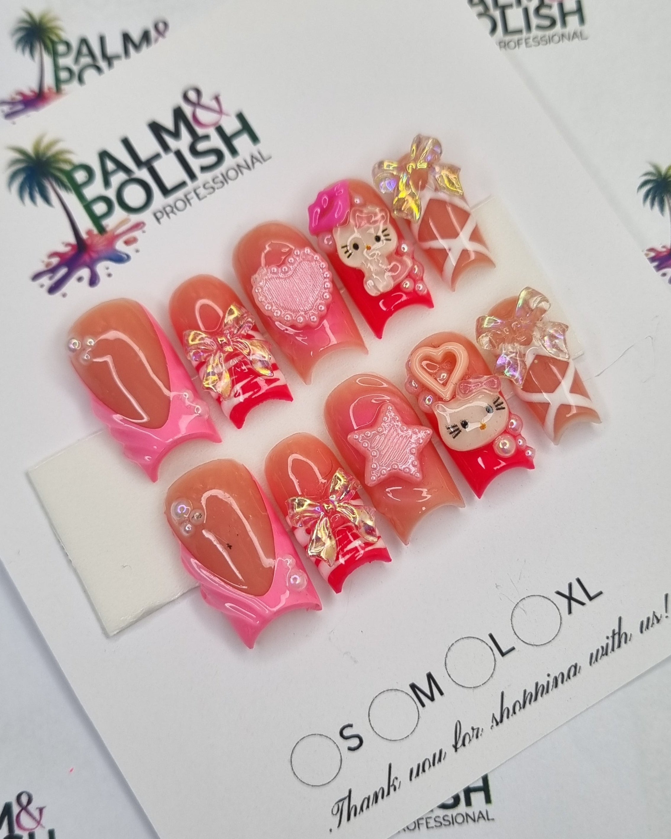 A set of HELLO KITY press-on nails displayed on a white card, photographed from above on a branded white surface. The nails feature pink, red, and nude tones with glittery gold details, floral motifs, and some clear embellishments.