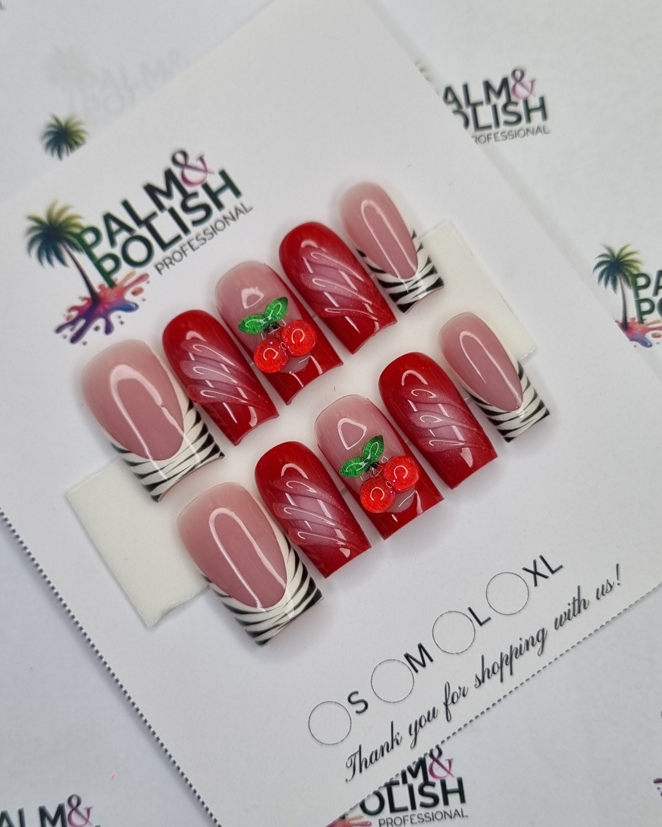 A set of 10 press-on nails displayed on a branded card, featuring red and pink designs with white stripes, cherries, and green leaves, set against a white patterned background.