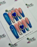 A set of press-on nails displayed on a white card, featuring blue and pink gradient nails with leopard print accents, rhinestone details, and two blue heart-shaped gems with rhinestone borders. The card is placed on a white background printed with the Palm & Polish logo presson nail vendor. presson nail factory.
