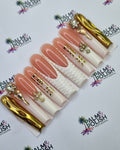 A set of gold acrylic press-on nails displayed on a white branded card. The nails feature a mix of nude pink, textured white, and metallic gold designs, with some nails decorated with rhinestones.
