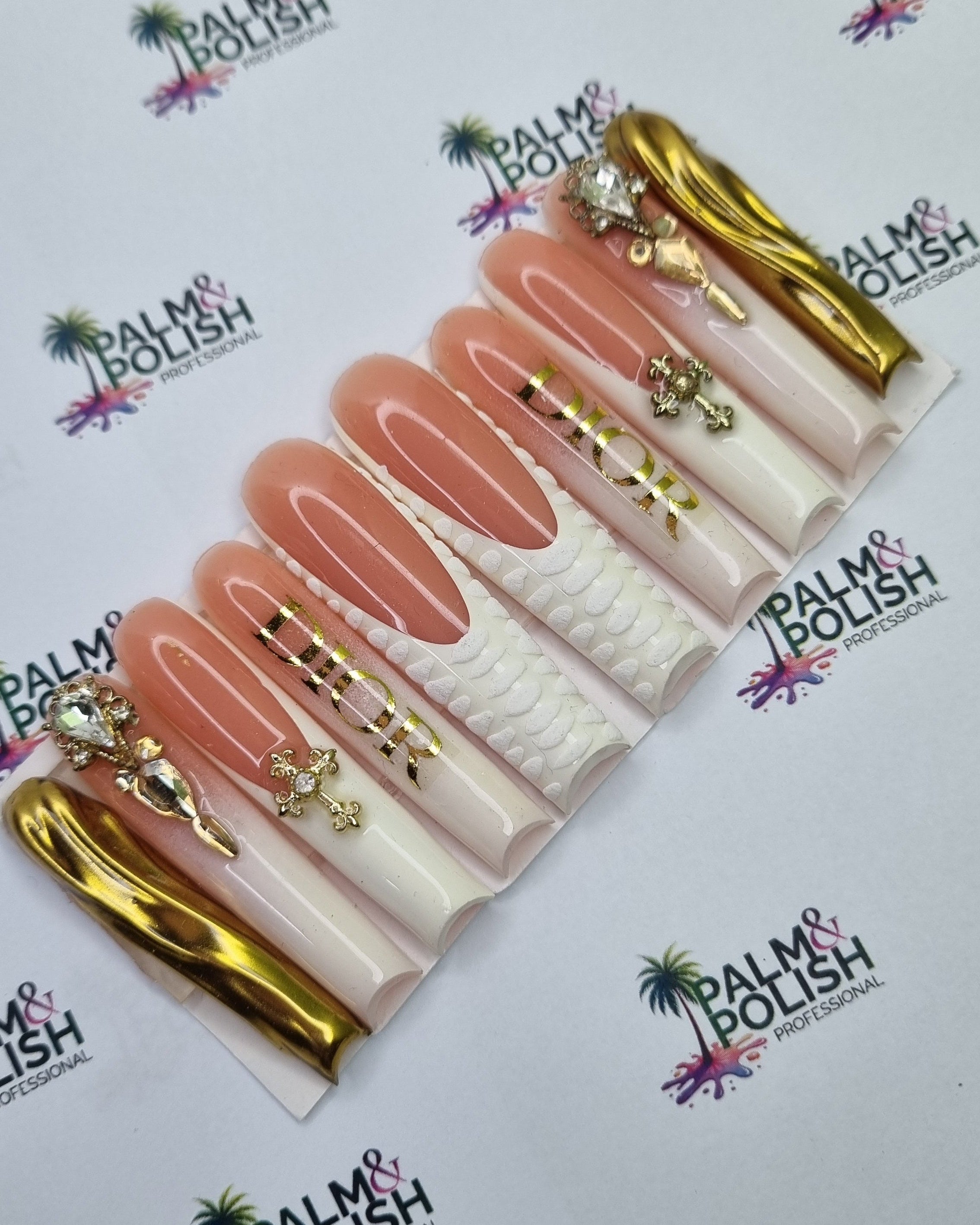 A set of gold acrylic press-on nails displayed on a white branded card. The nails feature a mix of nude pink, textured white, and metallic gold designs, with some nails decorated with rhinestones.
