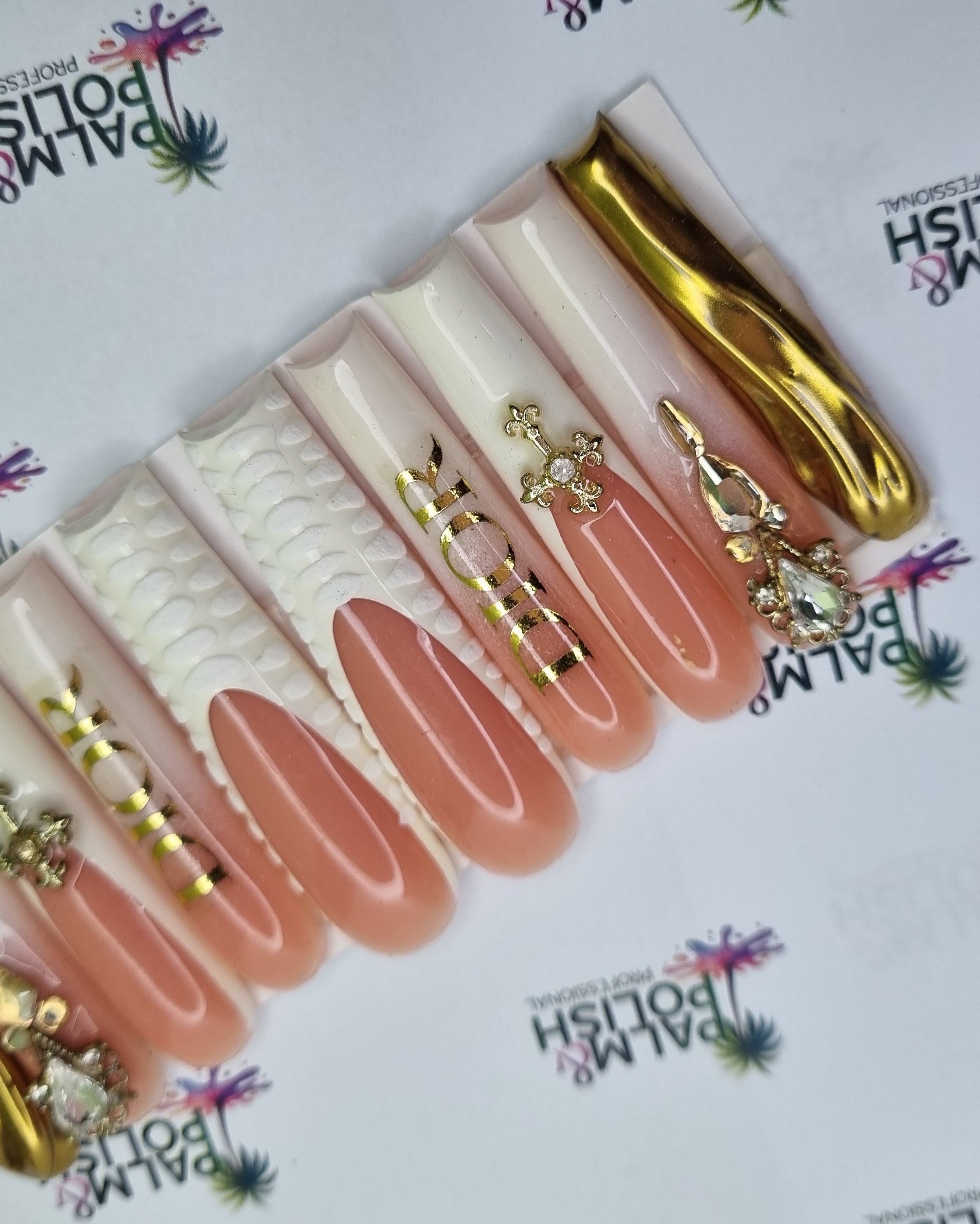 A set of false press-on nails displayed on a white branded card. The nails feature a mix of glossy nude pink, textured cream, and metallic gold designs, with one gold nail showing a floral pattern.