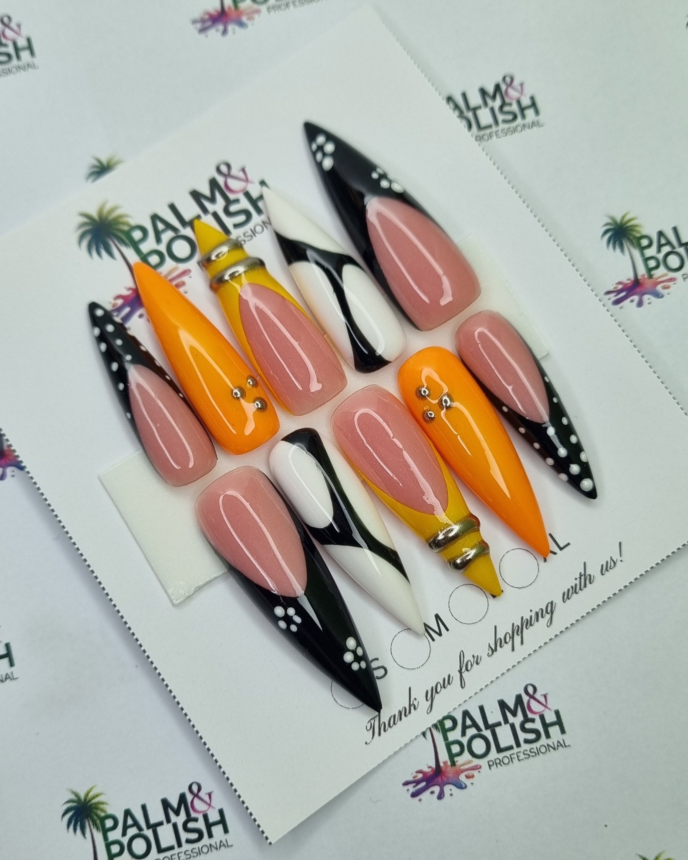 A set of artificial acrylic press-on nails displayed on a white backing card. The nails feature a mix of glossy pink, bright orange, black, and white designs, including some with small gold rhinestone accents.