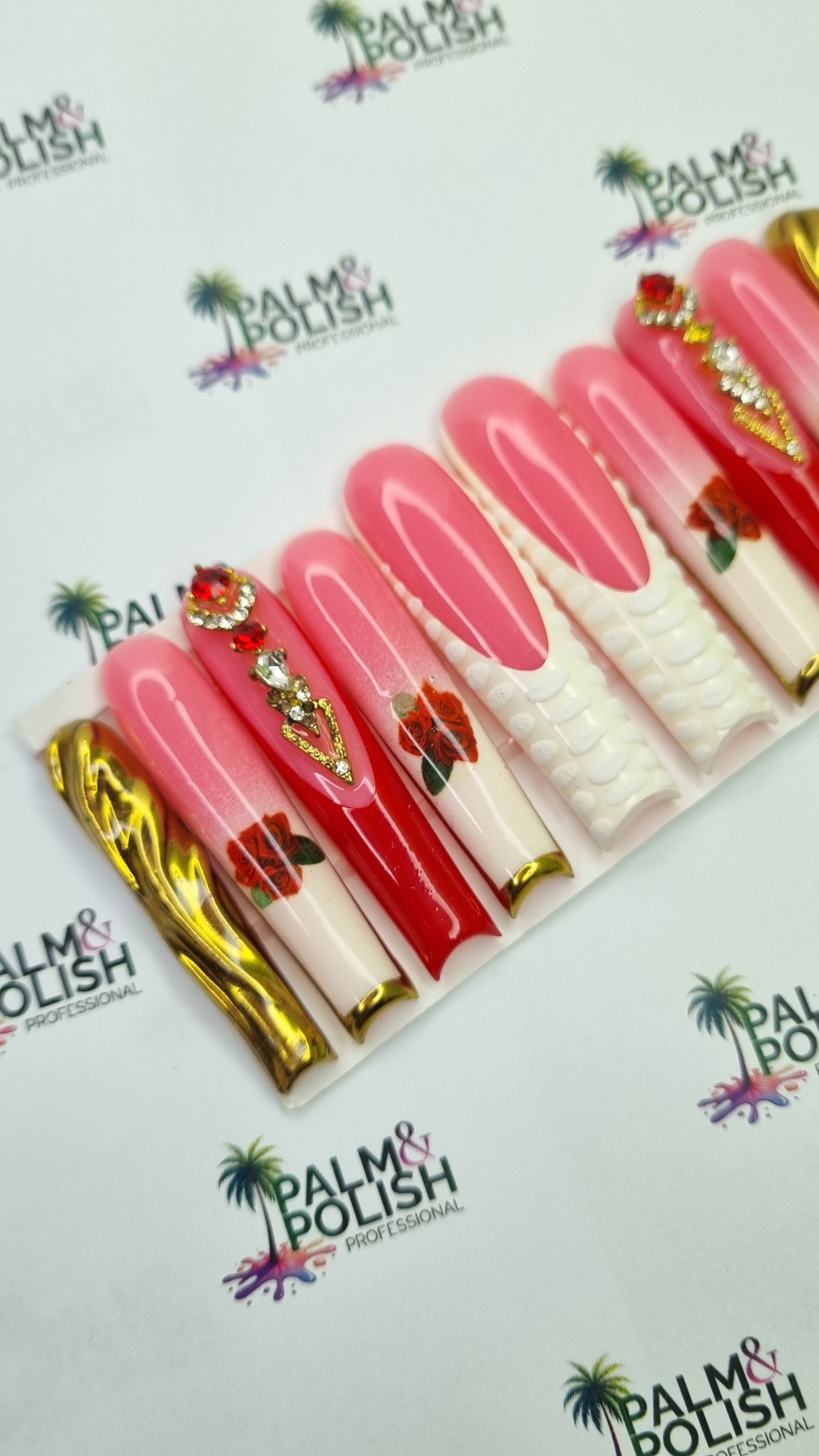 A set of artificial press-on nails displayed in a clear plastic tray, featuring pink, red, and white designs with gold foil, rhinestones, and floral details, set against a white branded background.