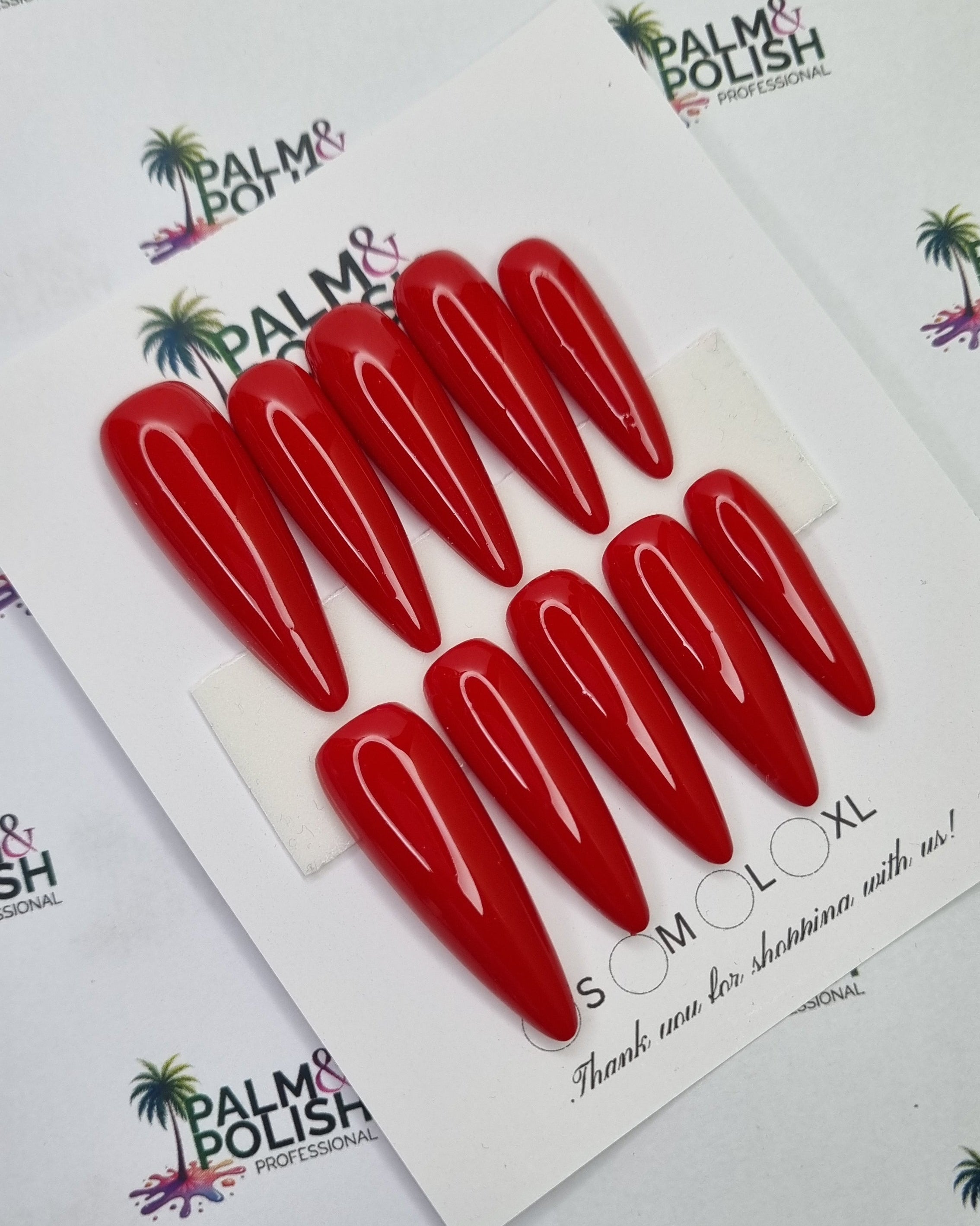 Six glossy red 2xl acrylic presson nail arranged on a white display card, photographed from above on a white branded surface with repeated Palm & Polish logos.