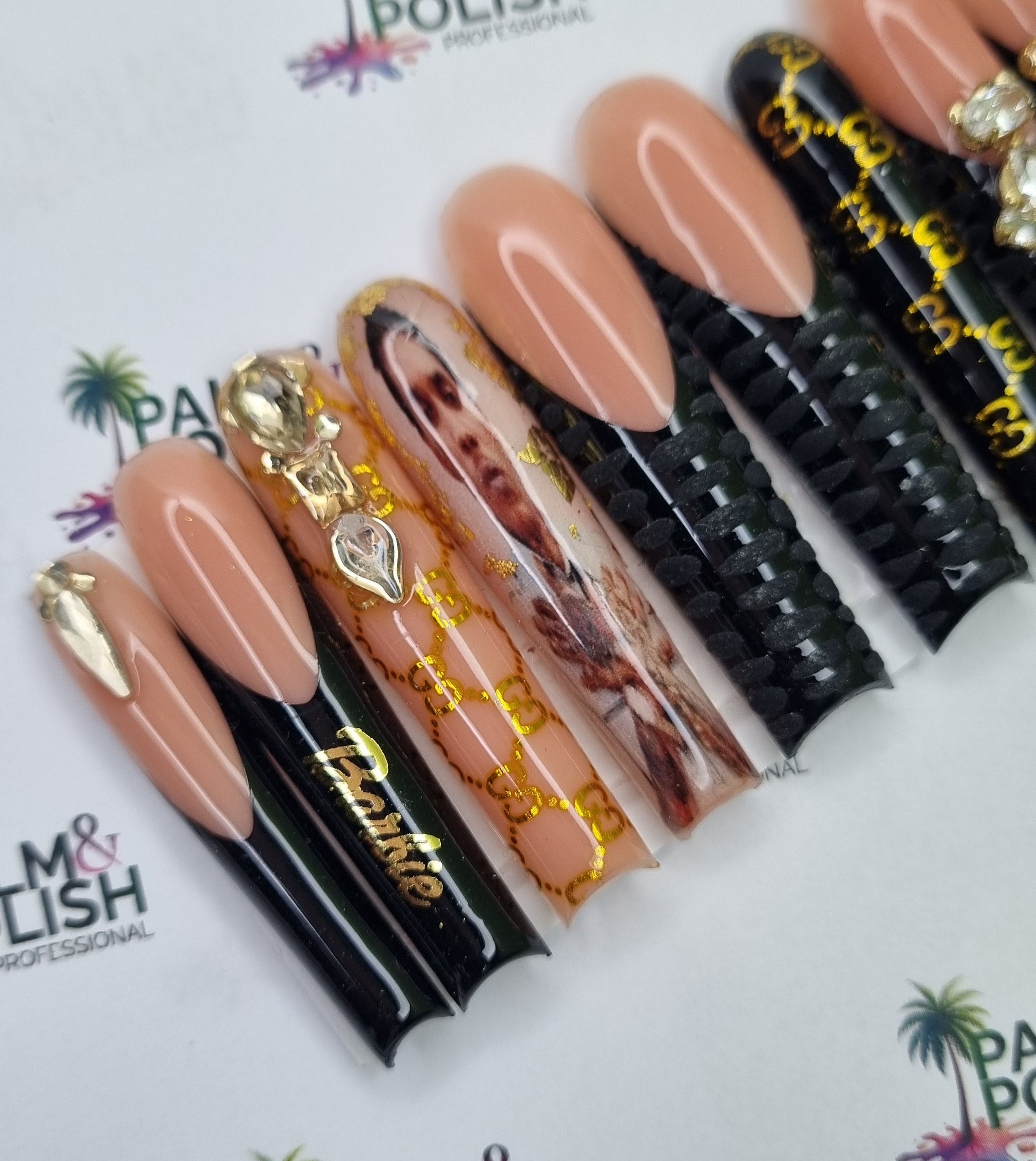 A set of artificial press-on nails displayed on black plastic forms. The nails feature a mix of solid nude pink, glossy black with gold foil script, clear with gold foil chains, and a glossy nail with a printed portrait of vybz kartel